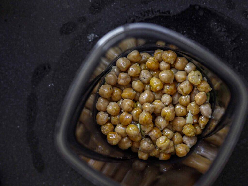 A top view of chickpeas inside a blender jar on a dark surface. The chickpeas are light brown, some glistening with moisture, and are ready for blending.